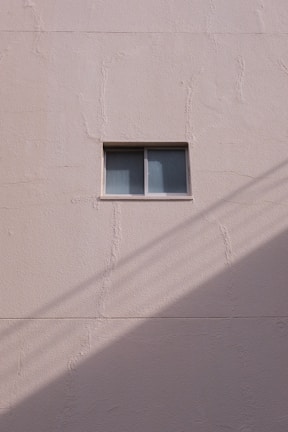 A simple beige wall with a small rectangular window, featuring a sliding design. The wall's texture appears rough and paint has subtle cracks. A shadow, likely from a utility line or similar object, diagonally crosses the bottom section of the image.