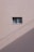 A simple beige wall with a small rectangular window, featuring a sliding design. The wall's texture appears rough and paint has subtle cracks. A shadow, likely from a utility line or similar object, diagonally crosses the bottom section of the image.