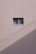 A simple beige wall with a small rectangular window, featuring a sliding design. The wall's texture appears rough and paint has subtle cracks. A shadow, likely from a utility line or similar object, diagonally crosses the bottom section of the image.