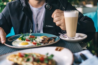 A table setting features a breakfast spread with a plate of sunny-side-up eggs garnished with herbs and cherry tomatoes. A glass of layered coffee with foam sits next to it on a saucer, and a person in casual attire holds a camera or similar device.