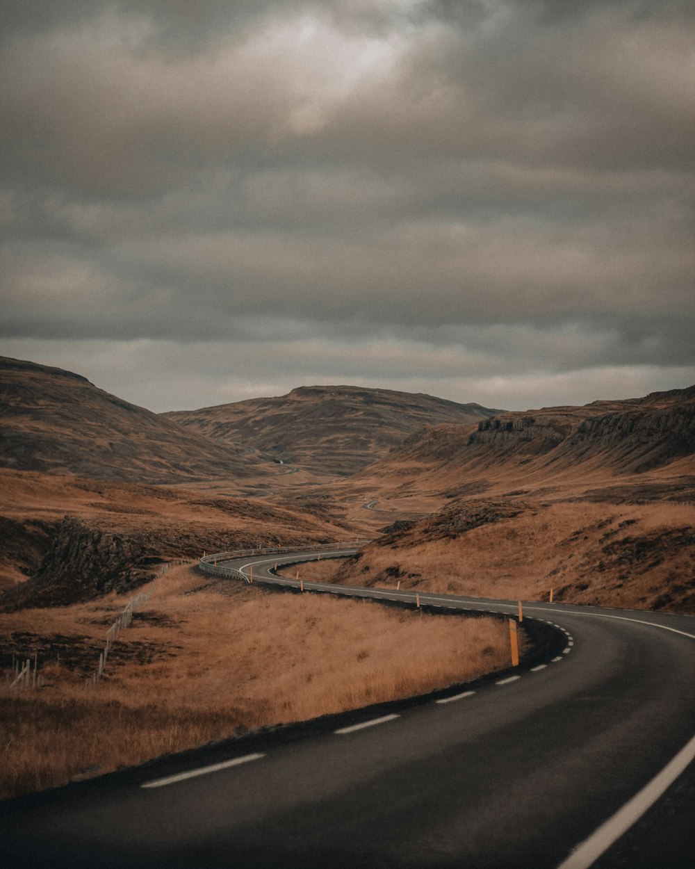 black asphalt road between brown mountains under gray cloudy sky during daytime