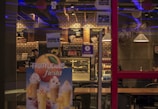 The image depicts the interior of a modern cafe with dim lighting featuring geometric wall designs, hanging lamps, and wooden tables. Menu boards are displayed on the wall, showcasing various food options. A refrigerated display filled with assorted beverages and sweets is visible, and there is a promotional poster for fruity desserts in the foreground.