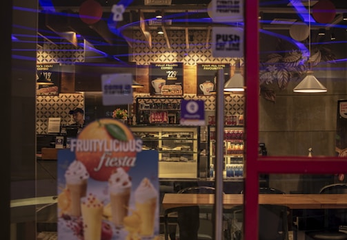 The image depicts the interior of a modern cafe with dim lighting featuring geometric wall designs, hanging lamps, and wooden tables. Menu boards are displayed on the wall, showcasing various food options. A refrigerated display filled with assorted beverages and sweets is visible, and there is a promotional poster for fruity desserts in the foreground.