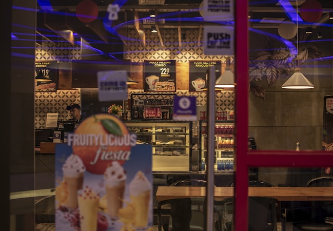 The image depicts the interior of a modern cafe with dim lighting featuring geometric wall designs, hanging lamps, and wooden tables. Menu boards are displayed on the wall, showcasing various food options. A refrigerated display filled with assorted beverages and sweets is visible, and there is a promotional poster for fruity desserts in the foreground.