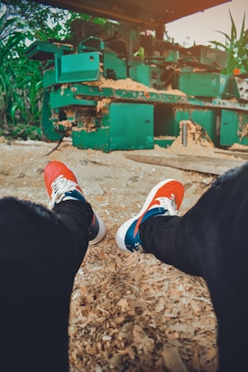 A person is seated on the ground covered with wood shavings, wearing red and white sneakers. In front, there is a large, industrial green machine, possibly used for wood processing. Surrounding the machine are plants and signs of an outdoor environment.