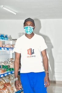 A person wearing a mask and cap stands in a well-lit retail store with shelves stocked with various packaged goods.