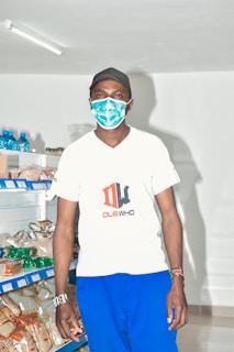 A person wearing a mask and cap stands in a well-lit retail store with shelves stocked with various packaged goods.
