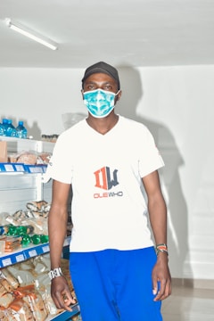 A person wearing a mask and cap stands in a well-lit retail store with shelves stocked with various packaged goods.