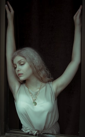 A woman with closed eyes and arms raised above her head, wearing a sleeveless dress and a gold necklace, stands with a serene expression. Her light hair contrasts with the dark background.