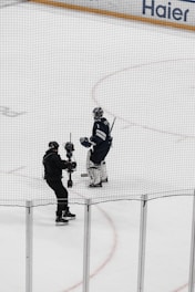 A hockey player in full gear stands on the ice next to a person holding a camera on a stabilizing mount. Both individuals are on an indoor hockey rink surrounded by safety nets.