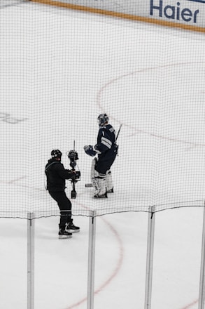 A hockey player in full gear stands on the ice next to a person holding a camera on a stabilizing mount. Both individuals are on an indoor hockey rink surrounded by safety nets.
