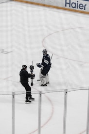 A hockey player in full gear stands on the ice next to a person holding a camera on a stabilizing mount. Both individuals are on an indoor hockey rink surrounded by safety nets.