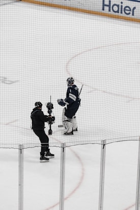 A hockey player in full gear stands on the ice next to a person holding a camera on a stabilizing mount. Both individuals are on an indoor hockey rink surrounded by safety nets.