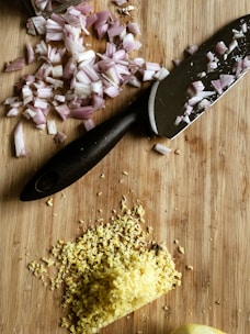 A wooden cutting board with chopped red onions and grated ginger next to a black-handled kitchen knife.