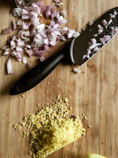 A wooden cutting board with chopped red onions and grated ginger next to a black-handled kitchen knife.