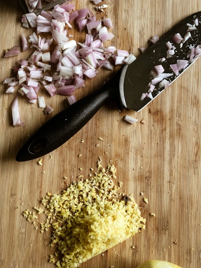 A wooden cutting board with chopped red onions and grated ginger next to a black-handled kitchen knife.