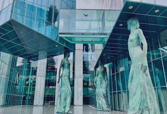 Three large statues of women in flowing robes stand in an atrium of a modern, glass-facade building. The reflective surfaces create an interplay of light and shadows, adding to the architectural aesthetics.