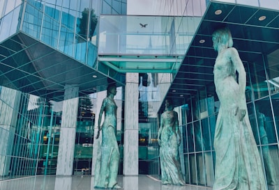 Three large statues of women in flowing robes stand in an atrium of a modern, glass-facade building. The reflective surfaces create an interplay of light and shadows, adding to the architectural aesthetics.