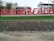 Large red letters spelling 'ALIBAG BEACH' are prominently displayed on a manicured grass platform. The backdrop consists of modern white buildings with red accents surrounded by palm trees. The foreground features a patterned array of concrete structures on a sandy surface.