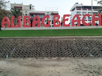 Large red letters spelling 'ALIBAG BEACH' are prominently displayed on a manicured grass platform. The backdrop consists of modern white buildings with red accents surrounded by palm trees. The foreground features a patterned array of concrete structures on a sandy surface.