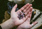 A close-up view of an open hand holding several polished semi-precious stones with purple hues. The stones have various shapes and some feature engraved symbols. The background is blurred and includes items like fabric and bottles.