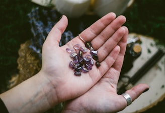 A close-up view of an open hand holding several polished semi-precious stones with purple hues. The stones have various shapes and some feature engraved symbols. The background is blurred and includes items like fabric and bottles.