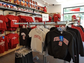 A retail display in a store features Stanford University merchandise, including red and grey t-shirts, hoodies, and sports equipment like basketballs and footballs on shelves and racks.