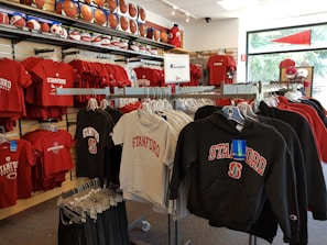 A retail display in a store features Stanford University merchandise, including red and grey t-shirts, hoodies, and sports equipment like basketballs and footballs on shelves and racks.