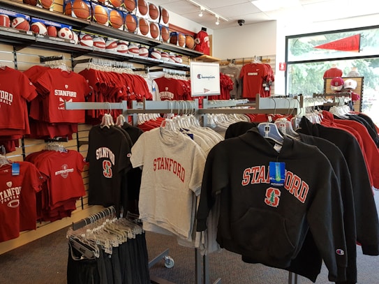 A retail display in a store features Stanford University merchandise, including red and grey t-shirts, hoodies, and sports equipment like basketballs and footballs on shelves and racks.
