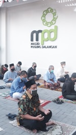 A group of people is seated in an outdoor area, each on a prayer mat, engaged in prayer. They are socially distanced and wearing masks. A sign with the words 'Masjid Pogung Dalangan' is visible on the wall behind them. The attire includes traditional clothing, indicating a cultural or religious gathering.