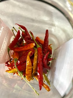 A collection of vibrant red and yellow chili peppers are gathered together inside a clear glass container. The peppers are variously shaped and sized, with smooth skin and green stems still attached. The lighting highlights their glossy texture, creating a lively and colorful display.