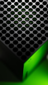 A close-up view of a device with a perforated surface featuring circular holes. The structure has a gradient of green light illuminating a portion of it, suggesting a modern and sleek design.
