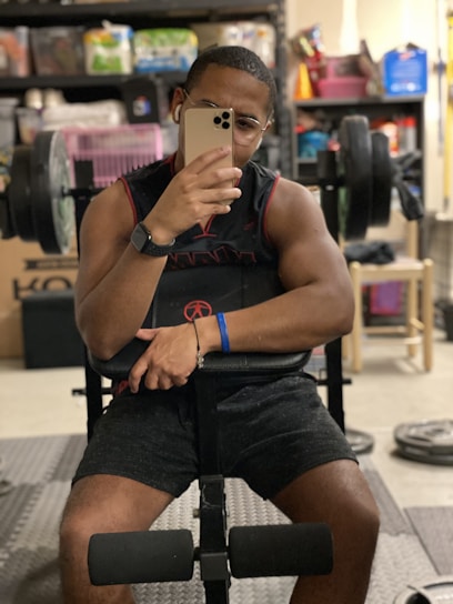 A person sitting on a weight bench in what appears to be a home gym. They are taking a selfie using a smartphone and wearing glasses, a sleeveless top, shorts, and a wristband. Behind them are various fitness equipment, a storage rack with household items, and a pink pet carrier. The environment appears organized and functional.
