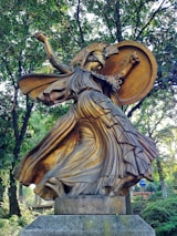 A bronze statue of a warrior woman with a helmet adorned with a crest, holding a large round shield. Her flowing robes swirl dramatically around her, giving a sense of motion. The sculpture is situated in a wooded area, surrounded by lush green trees.