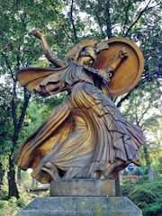 A bronze statue of a warrior woman with a helmet adorned with a crest, holding a large round shield. Her flowing robes swirl dramatically around her, giving a sense of motion. The sculpture is situated in a wooded area, surrounded by lush green trees.