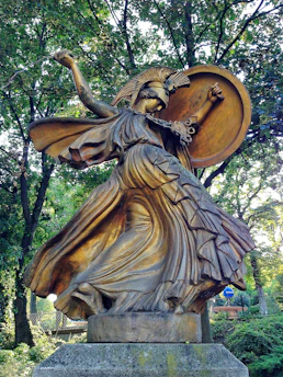 A bronze statue of a warrior woman with a helmet adorned with a crest, holding a large round shield. Her flowing robes swirl dramatically around her, giving a sense of motion. The sculpture is situated in a wooded area, surrounded by lush green trees.