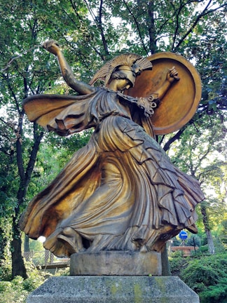A bronze statue of a warrior woman with a helmet adorned with a crest, holding a large round shield. Her flowing robes swirl dramatically around her, giving a sense of motion. The sculpture is situated in a wooded area, surrounded by lush green trees.