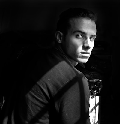 A black and white portrait of a man with a serious expression, seated and slightly turned to the side, with light and shadow creating dramatic contrasts on his face.