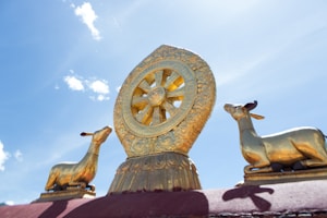 A golden Dharmachakra wheel flanked by two golden deer statues on a pedestal, set against a clear blue sky with a few wispy clouds.