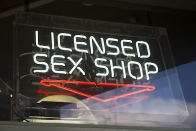 A neon sign with the words 'LICENSED SEX SHOP' written in white, situated inside a transparent rectangle frame that appears to be hanging in a window. Below the main text, there are red neon shapes. The sign is reflective, showing hints of the surroundings outside the window.