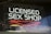 A neon sign with the words 'LICENSED SEX SHOP' written in white, situated inside a transparent rectangle frame that appears to be hanging in a window. Below the main text, there are red neon shapes. The sign is reflective, showing hints of the surroundings outside the window.