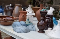 A collection of various ceramic and earthenware pots and vases are arranged on a white table. The vessels are of different shapes and sizes, ranging from small, intricately designed pieces to larger, more simplistic ones. Colors vary, including shades of brown, white, blue, and teal, displaying a mix of glazed and matte finishes. The background features a stone wall and some greenery, adding a rustic outdoor setting.