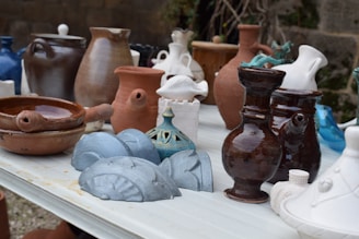 A collection of various ceramic and earthenware pots and vases are arranged on a white table. The vessels are of different shapes and sizes, ranging from small, intricately designed pieces to larger, more simplistic ones. Colors vary, including shades of brown, white, blue, and teal, displaying a mix of glazed and matte finishes. The background features a stone wall and some greenery, adding a rustic outdoor setting.