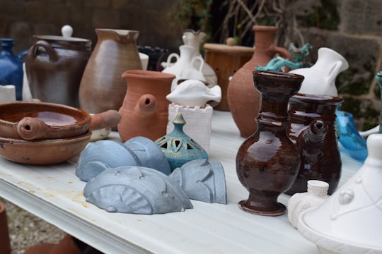 A collection of various ceramic and earthenware pots and vases are arranged on a white table. The vessels are of different shapes and sizes, ranging from small, intricately designed pieces to larger, more simplistic ones. Colors vary, including shades of brown, white, blue, and teal, displaying a mix of glazed and matte finishes. The background features a stone wall and some greenery, adding a rustic outdoor setting.