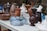 A collection of various ceramic and earthenware pots and vases are arranged on a white table. The vessels are of different shapes and sizes, ranging from small, intricately designed pieces to larger, more simplistic ones. Colors vary, including shades of brown, white, blue, and teal, displaying a mix of glazed and matte finishes. The background features a stone wall and some greenery, adding a rustic outdoor setting.