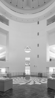 A spacious, modern, and minimalist interior with a large central area featuring geometric patterned flooring. The room is surrounded by multiple levels of balconies and glass display cases. The walls are bright and predominantly feature a circular architectural design. There are a couple of people standing on the right balcony.