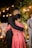 A couple is embracing, with the man wearing a dark suit and the woman in a red dress with embroidered patterns. Soft, warm lighting creates a romantic atmosphere with visible string lights strung in the background.