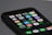 A smartphone displaying its home screen with various colorful app icons including calendar, photos, maps, weather, clock, and more. The phone is placed on a dark surface at an angle.