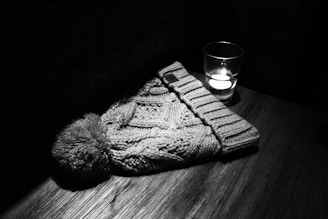 A knitted beanie hat with a pom-pom is placed on a wooden surface beside a lit candle in a glass holder, creating a cozy and intimate scene.