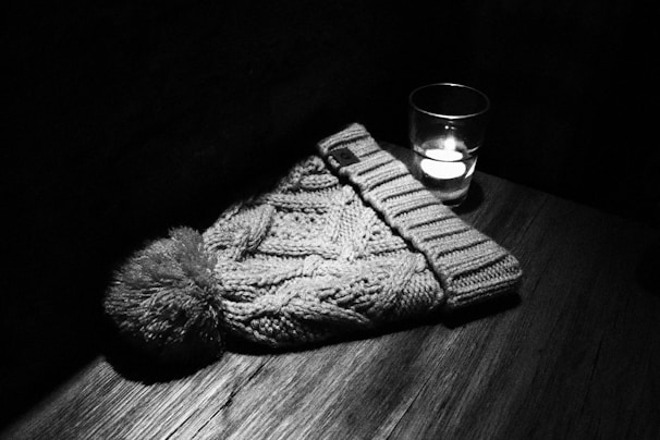 A knitted beanie hat with a pom-pom is placed on a wooden surface beside a lit candle in a glass holder, creating a cozy and intimate scene.